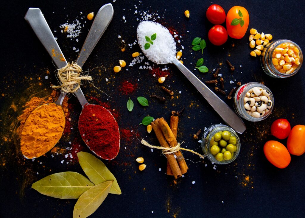 Top view of dry turmeric and paprika near bay leaves with cinnamon sticks and sea salt in spoons near jars with popcorn grains and olives with ripe colorful cherry tomatoes
